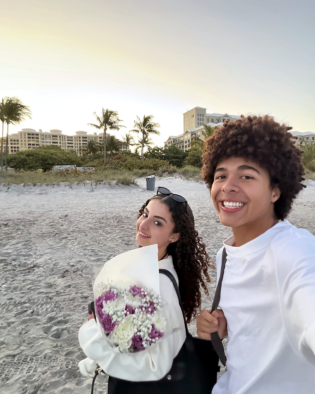 Elena at the beach holding a bouquet of purple and white flowers.