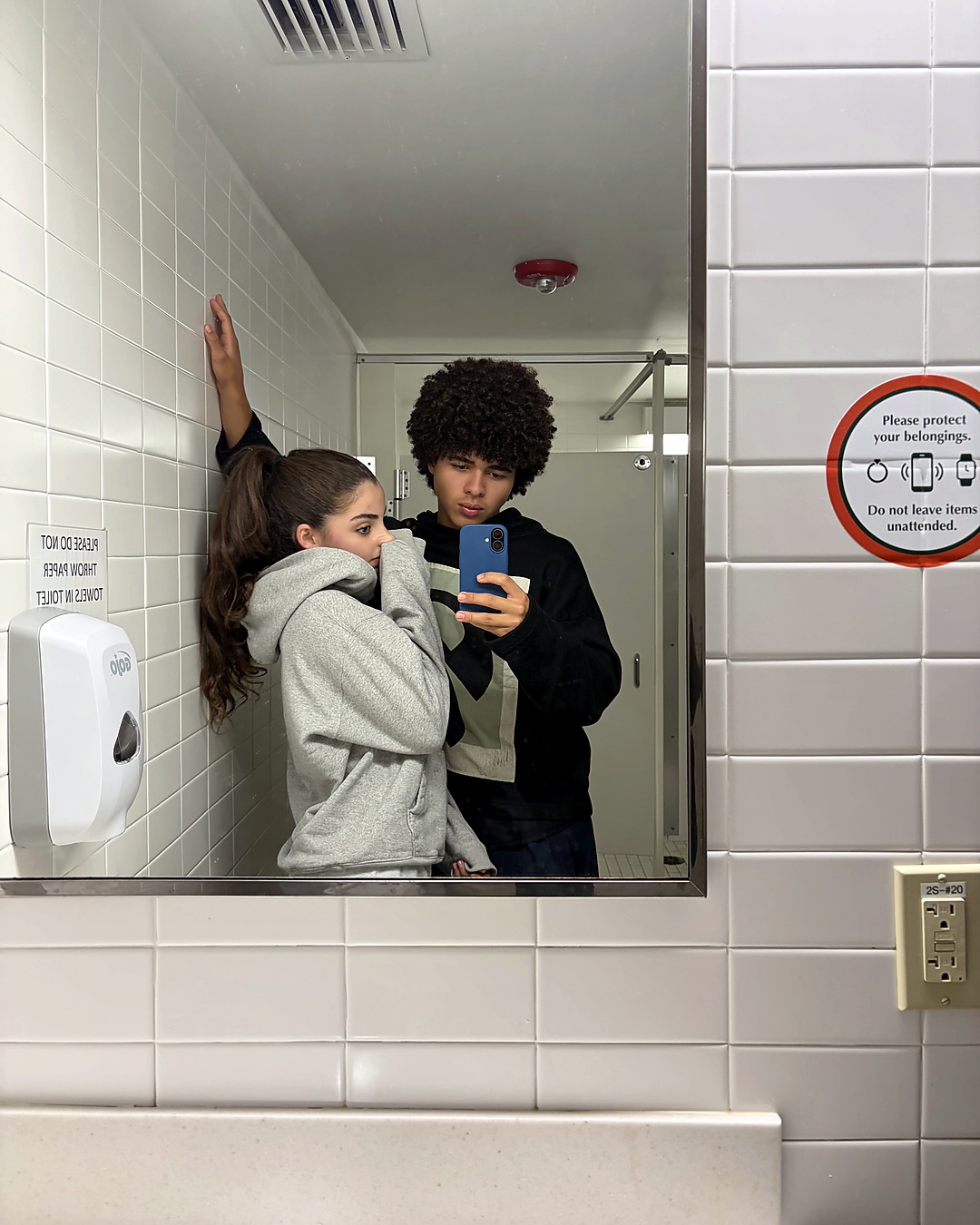 A school bathroom mirror, Elena in a grey hoodie covering her face.